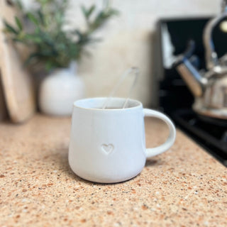 Tea cup featuring heart-shaped design, infuser nestled inside, set on counter alongside teapot.